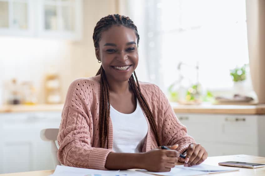 Teenage girl with natural hair smiling at the camera while doing her homework in the kitchen