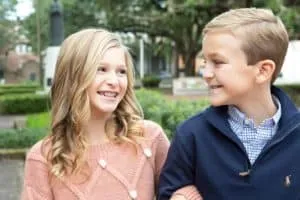 A blond brother and sister linking arms and smiling at each other in a park