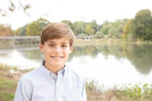 A boy with brown hair and braces smiling in front of a lake