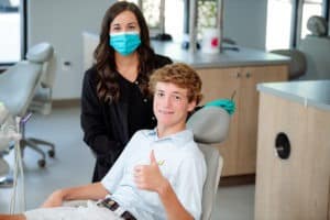 Female orthodontist with a blue mask and a boy with brace in a treatment chair giving a thumbs up