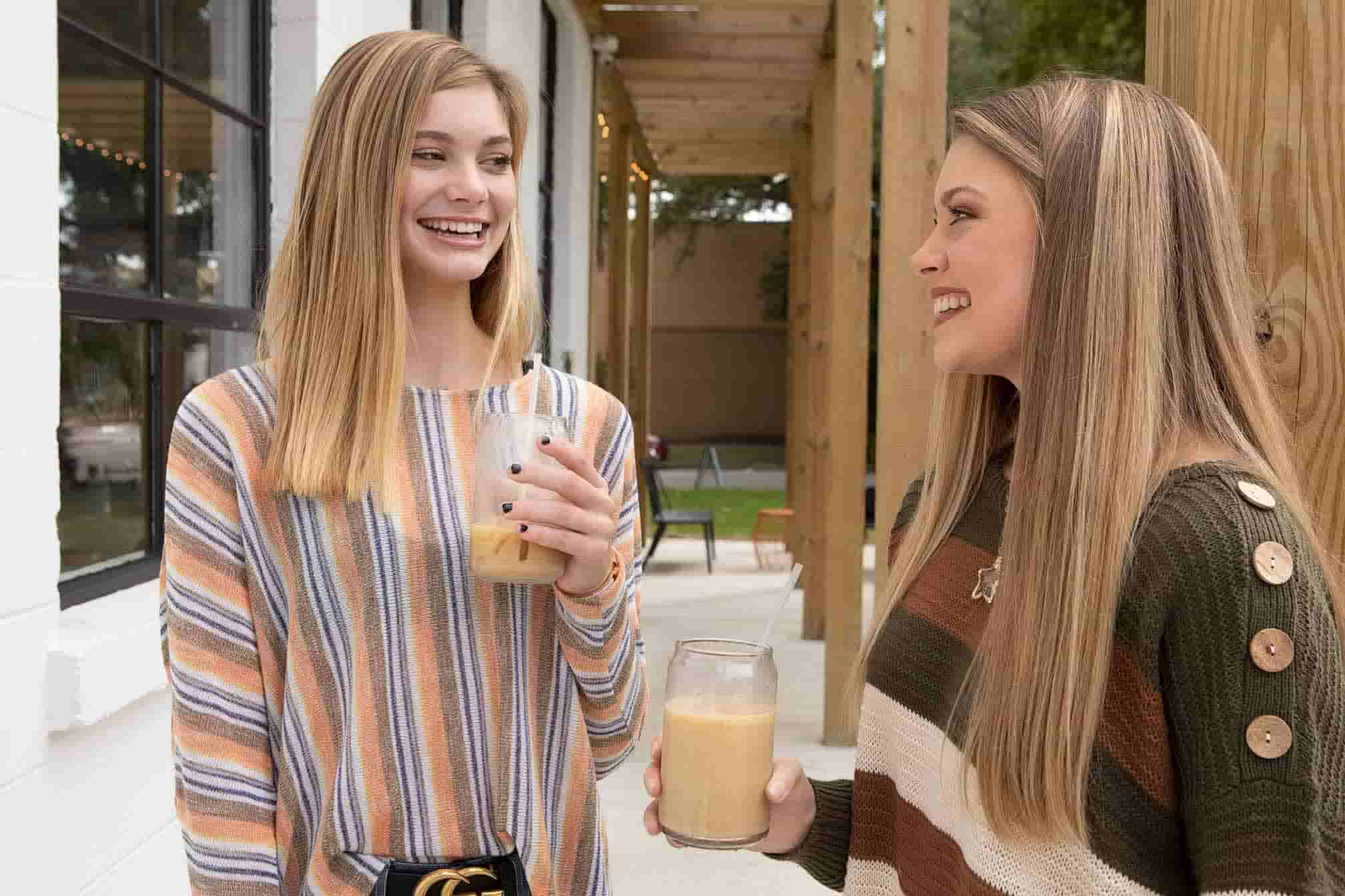 Two girls with straight blond hair standing under a pergola and drinking iced coffee